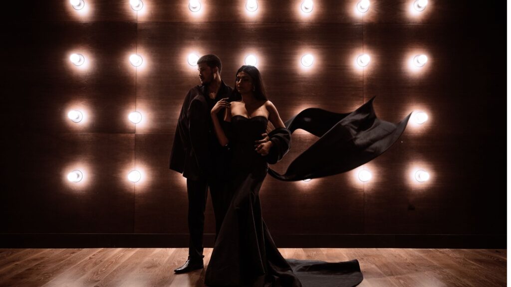 A dramatic and moody portrait of a couple standing in front of a wall adorned with evenly spaced light bulbs. The woman, wearing a strapless black gown with a long, flowing train, is standing confidently as her dress billows in the air. The man, dressed in dark attire, stands close beside her, gazing in the opposite direction. The warm lighting creates a striking contrast against the dark, elegant outfits, adding depth and intensity to the scene.